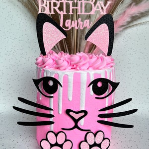 Puede incluir: Pastel de temática de gato rosa y negro con glaseado blanco y un adorno de "Feliz cumpleaños Laura".