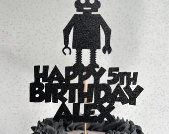 May include: Black glitter cake topper with a robot design and the text "Happy 5th Birthday Alex".