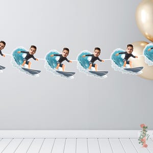 May include: A decorative banner with surfer cutouts. Each cutout shows a person surfing a wave. The person wears a black wetsuit and rides a grey surfboard. The background includes gold balloons and a white string.