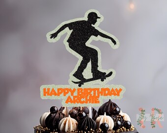 May include: A black glitter skateboarder cake topper with the text "HAPPY BIRTHDAY ARCHIE" in orange. The topper is placed on top of a chocolate cake decorated with white and dark chocolate candies.