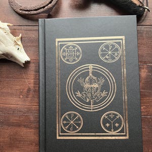 Könnte beinhalten: Ein schwarzes Hardcover-Buch mit einem goldgeprägten Design. Das Design zeigt einen Frosch in einem Kreis, umgeben von vier kleineren Kreisen mit Symbolen. Das Buch hat einen strukturierten Einband.