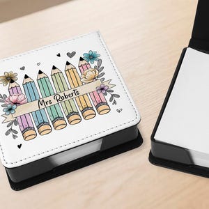 Könnte beinhalten: Ein schwarzer Notizblockhalter mit weißem Deckel, der ein Bleistiftdesign und den Text "Mrs Roberts" zeigt. Der Notizblockhalter ist geöffnet und zeigt einen Stapel weißes Papier. Der Notizblock liegt auf einer Holzoberfläche.