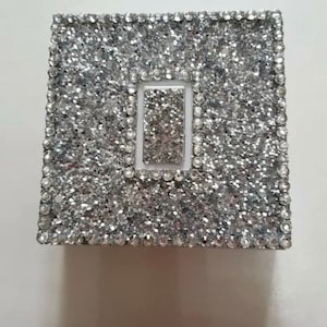 May include: A silver glitter light switch cover with a white inner frame. The cover is decorated with small silver glitter and rhinestones.
