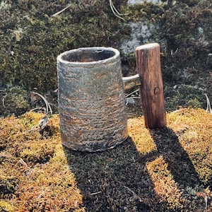 May include: A handcrafted, textured metal mug with a wooden handle. The mug has a cylindrical shape and a rough, metallic finish. The handle is made of a dark, polished wood. The mug sits on a bed of green and brown moss.