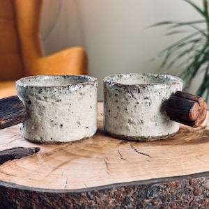 May include: Two handmade ceramic mugs with speckled, off-white exteriors and dark brown wooden handles. The mugs are displayed on a rustic wooden surface, showcasing a natural, earthy aesthetic. The mugs are perfect for coffee or tea.