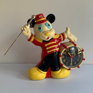 May include: A vintage Mickey Mouse figurine clock. The figurine is dressed in a red and black band uniform, holding a baton and a drum-shaped clock. The clock face is black with white numbers and red hands. The figurine has yellow shoes.