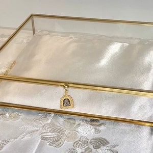 May include: A rectangular glass and gold-toned metal display box. The box has a hinged lid and a small decorative charm. The interior is lined with white fabric. The box is designed for displaying jewelry or small items.