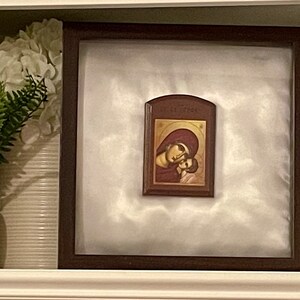 May include: A framed religious icon depicting a mother and child, set against a white background. The icon features warm tones of gold, red, and brown. The frame is dark wood, and the image is displayed on a shelf with decorative elements.