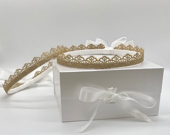 May include: A white rectangular gift box featuring a white satin ribbon bow on the front. Resting on top of the box is a circular headband adorned with intricate gold lace trim, with an additional length of the same gold lace trim extending outward.