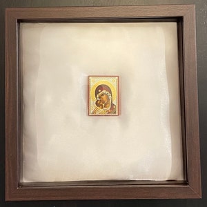 May include: A framed religious icon featuring a depiction of the Virgin Mary and the Christ Child. The icon is set against a white background within a dark brown wooden frame. The icon's colors include gold, red, and brown.