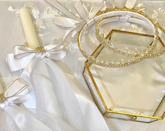 May include: Ivory-colored candles adorned with white satin bows and gold embellishments. Two jeweled crowns rest on a gold-framed, hexagonal glass box. The items are arranged on a white, floral-patterned fabric.