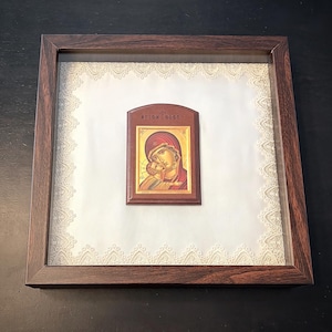 May include: A framed religious icon featuring a depiction of Mary and Jesus. The icon is set against a white background with a lace-like border and encased in a dark brown wooden frame. The icon itself is a rectangular shape with gold, red, and brown tones.
