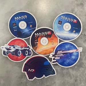 以下が含まれることがあります： 「Mass Effect」ビデオゲームシリーズのデザインが施された丸いステッカーのコレクション。ステッカーにはゲームタイトル、宇宙船、N7ロゴが含まれており、一部にはPlayStationのロゴも含まれています。