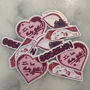 May include: A selection of stickers in pink and burgundy. The stickers display the text "I'm Your Freaky Nikki!" in a heart shape, the word "Obsession", and "One Wish Willow". Other stickers depict an eye.