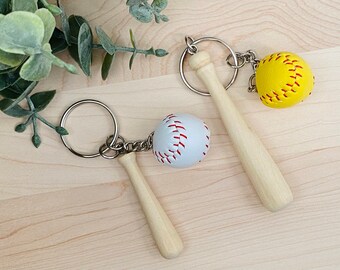 May include: Two baseball-themed keychains with wooden bats and baseballs. One keychain has a white baseball with red stitching and the other has a yellow softball with red stitching.