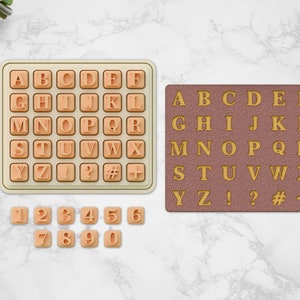 May include: A beige rectangular tray containing a grid of raised alphabet letters, punctuation marks, and symbols, alongside a separate set of individual beige number tiles and a brown rectangular mat featuring the same alphabet and symbols in gold.