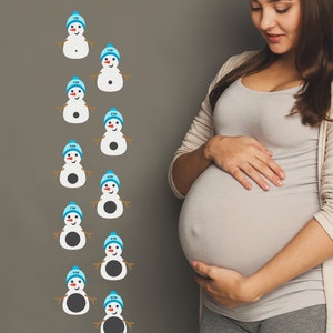 May include: A pregnant woman stands in front of a gray wall with a snowman chart showing the size of a baby at different stages of pregnancy. The chart says "Welcome to Frosty's Dilation Station".