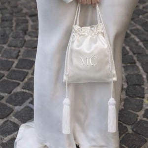 May include: Ivory satin drawstring bag with white rope handles and tassels. The bag features the embroidered initials "MC". The bag is held by a person wearing a white dress and silver sandals.