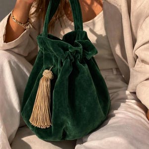 May include: A dark green velvet handbag with a drawstring closure and a gold tassel. The bag has a rounded shape and two short handles. The velvet material gives the bag a luxurious appearance.