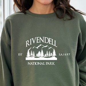 May include: A green sweatshirt with a white graphic of a mountain range and trees. The graphic says "Rivendell National Park" and "Est. S.A. 1697".