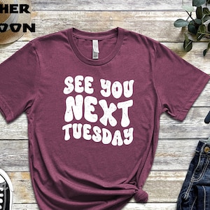 May include: A heather maroon t-shirt with the text "See You Next Tuesday" in white, retro-style lettering. The shirt is laid flat on a wooden surface with a pair of black sneakers, a watch, and a pair of jeans.