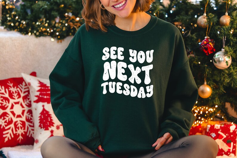 May include: A green sweatshirt with the text "See You Next Tuesday" in white, bubble letters. The sweatshirt is worn by a person sitting in front of a Christmas tree.