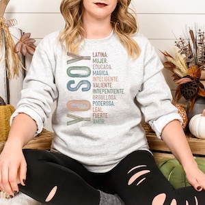 May include: A woman wearing a grey sweatshirt with the text "YO SOY" and a list of Spanish words in colorful lettering. The words include "Latina", "Mujer", "Educada", "Magica", "Inteligente", "Valiente", "Independiente", "Orgullosa", "Poderosa", "Leal", "Fuerte", and "Amor".