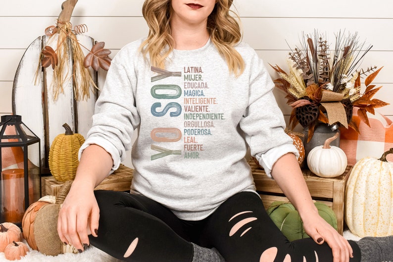 May include: A woman wearing a grey sweatshirt with the text "YO SOY" and a list of Spanish words in colorful lettering. The words include "Latina", "Mujer", "Educada", "Magica", "Inteligente", "Valiente", "Independiente", "Orgullosa", "Poderosa", "Leal", "Fuerte", and "Amor".