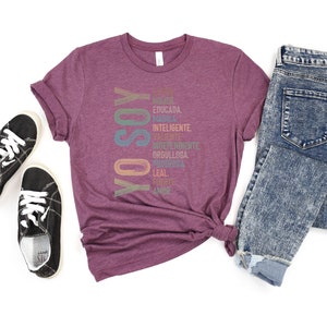 May include: A maroon t-shirt with the Spanish words "Yo Soy" printed vertically in rainbow colors. The words are surrounded by a list of Spanish words describing positive qualities, such as "mujer", "educada", "valiente", and "fuerte".