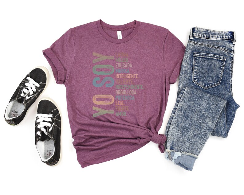 May include: A maroon t-shirt with the Spanish words "Yo Soy" printed vertically in rainbow colors. The words are surrounded by a list of Spanish words describing positive qualities, such as "mujer", "educada", "valiente", and "fuerte".