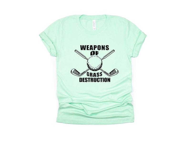 May include: A light green t-shirt with a black graphic of crossed golf clubs and a golf ball. The text "Weapons of Grass Destruction" is printed below the graphic.