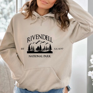 May include: A beige hoodie with a black graphic print of a mountain range and trees. The graphic says "Rivendell National Park" with the text "EST" and "S.A. 1697" on either side of the graphic.