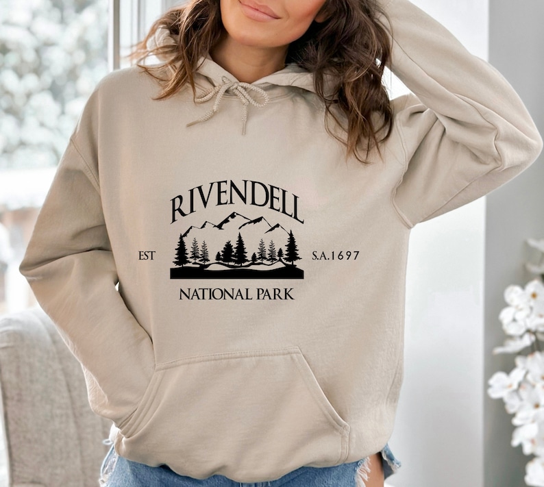 May include: A beige hoodie with a black graphic print of a mountain range and trees. The graphic says "Rivendell National Park" with the text "EST" and "S.A. 1697" on either side of the graphic.