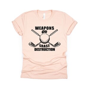 May include: A pink t-shirt with a black graphic that says "Weapons of Grass Destruction" with a golf ball and two crossed golf clubs.