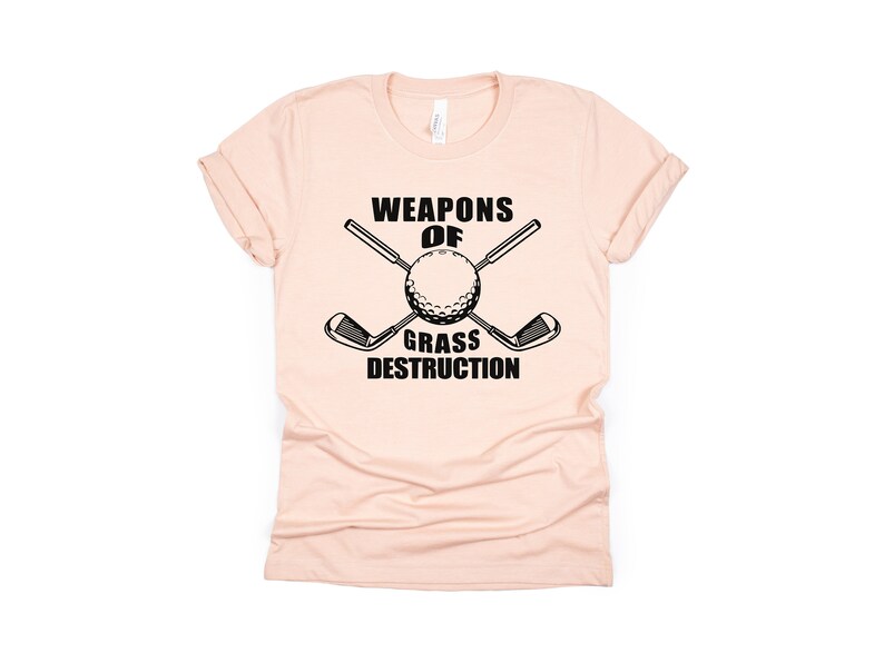 May include: A pink t-shirt with a black graphic that says "Weapons of Grass Destruction" with a golf ball and two crossed golf clubs.