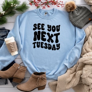May include: A light blue sweatshirt with the text "See You Next Tuesday" in black, retro-style lettering. The sweatshirt is laid out on a white surface with a pair of brown suede boots, a knitted scarf, and a gray beanie.