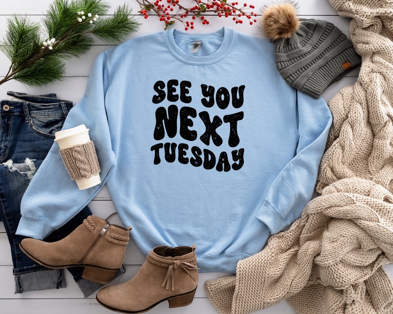 May include: A light blue sweatshirt with the text "See You Next Tuesday" in black, retro-style lettering. The sweatshirt is laid out on a white surface with a pair of brown suede boots, a knitted scarf, and a gray beanie.