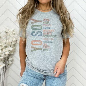 May include: A gray t-shirt with the Spanish words "YO SOY" in a vertical column, followed by a list of positive attributes in Spanish, including "Latina", "Mujer", "Educada", "Magica", "Inteligente", "Valiente", "Independiente", "Orgullosa", "Poderosa", "Leal", "Fuerte", and "Amor".