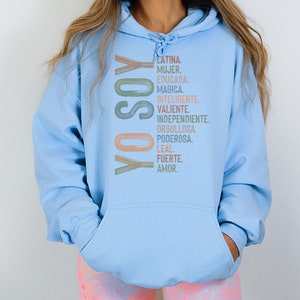May include: A light blue hoodie with the text "YO SOY" in a colorful, vertical design. The text is followed by a list of Spanish words describing positive qualities, including "Latina", "Mujer", "Educada", "Magica", "Inteligente", "Valiente", "Independiente", "Orgullosa", "Poderosa", "Leal", "Fuerte", and "Amor."