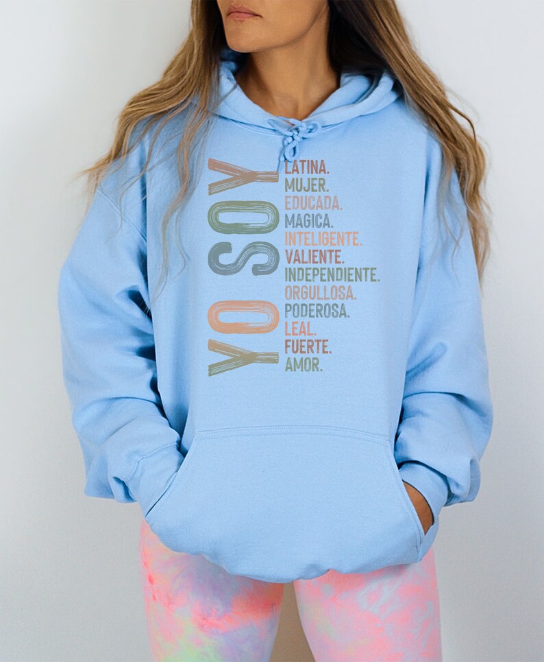 May include: A light blue hoodie with the text "YO SOY" in a colorful, vertical design. The text is followed by a list of Spanish words describing positive qualities, including "Latina", "Mujer", "Educada", "Magica", "Inteligente", "Valiente", "Independiente", "Orgullosa", "Poderosa", "Leal", "Fuerte", and "Amor."