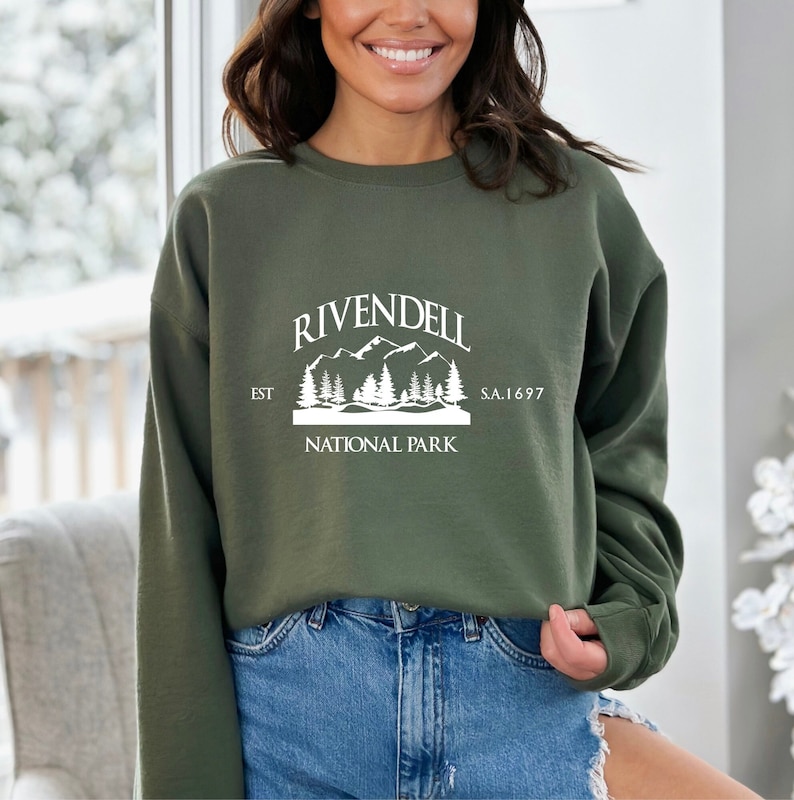 May include: A green sweatshirt with a white graphic of a mountain range and trees. The graphic says "Rivendell National Park" and "Est. S.A. 1697".