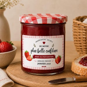 Peut inclure: Un pot en verre de confiture de fraises avec un couvercle à carreaux rouges et blancs. L'étiquette porte le texte "INGRÉDIENT SECRET DE NOTRE plus belle confiture BÉBÉ EN PRÉPARATION ARRIVÉE PRÉVUE JANVIER 2027" avec des illustrations de fraises.