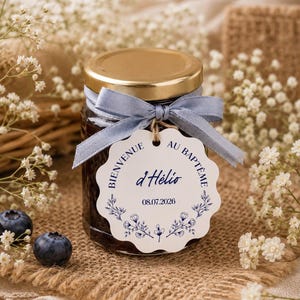 Peut inclure: Un petit pot en verre avec un couvercle doré, noué d'un ruban bleu clair et d'une étiquette blanche festonnée. L'étiquette présente des illustrations florales bleues et le texte : BIENVENUE AU BAPTÊME d'Hélio 08.07.2026. Contenu sombre visible.