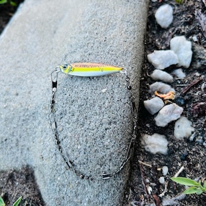May include: A yellow and silver fishing lure with a chain and a hook. The lure has a yellow body with brown and red markings and the number "7g" printed on it. The chain is made of small, dark metal links.
