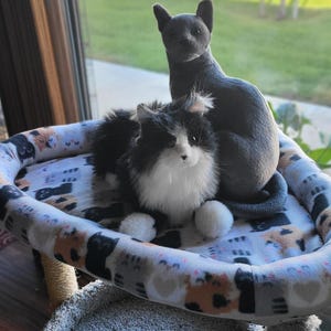 May include: A cat bed with a patterned design featuring various cat illustrations. Two plush cat toys, one gray and one black and white, are resting on the bed. The bed is oval-shaped and sits on a cat tree.