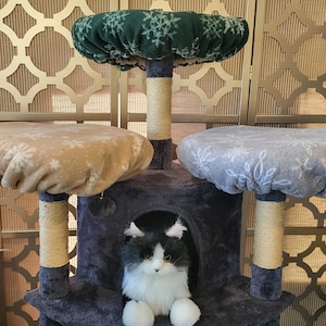 May include: A multi-level cat tree with three platforms and a hideaway. The platforms are covered in beige, gray, and green fabric with a snowflake pattern. The cat tree has sisal-wrapped posts and a plush, dark gray base. A black and white cat is inside the hideaway.