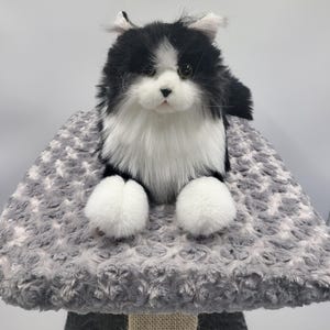 May include: A plush cat toy with black and white fur, perched on a gray, textured cat tree. The cat has white paws and a fluffy chest. The cat tree has a soft, patterned surface.