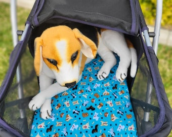 May include: A plush beagle dog toy with brown, white, and black fur resting inside a black pet stroller. The stroller features a bright blue interior liner decorated with a repeating pattern of small colorful dogs and white bone shapes.