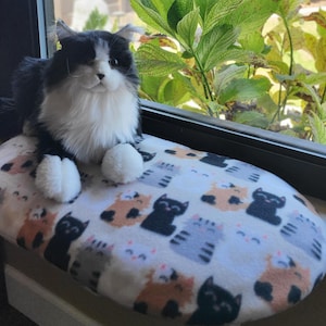 May include: A plush black and white cat toy sits on an oval-shaped cat window perch. The perch features a white fabric cover with a repeating pattern of small, colorful cartoon cats in shades of black, gray, orange, and pink on a white background.