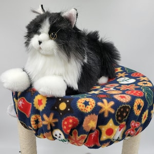 May include: A plush black and white cat toy with long fur, resting on a circular platform covered in a dark blue fleece fabric featuring a colorful pattern of various holiday cookies, including stars, circles, and snowman shapes.